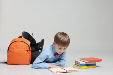 Sırt çantası açık gri arka planda kitap okuyan sevimli bir okul çocuğu.