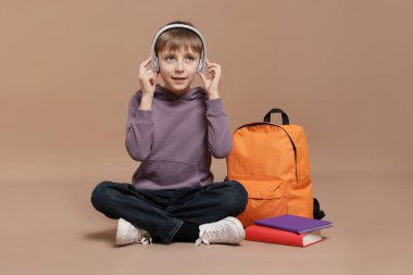 Sırt çantası ve kahverengi arka plan kitapları olan kulaklıklı tatlı bir okul çocuğu.