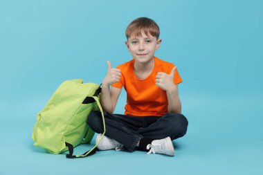 Sırt çantası açık mavi arka planda baş parmak gösteren sevimli okul çocuğu.