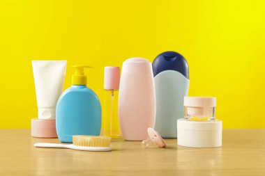 Baby cosmetic products and bath accessories on wooden table against yellow background
