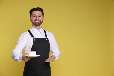 Sarı arka planda bir fincan sıcak içecekle gülümseyen baristanın portresi. Metin için boşluk