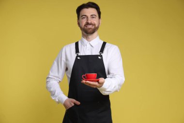 Sarı arka planda bir fincan sıcak içecekle gülümseyen baristanın portresi.