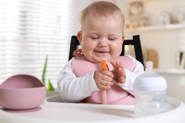 Evdeki beslenme sandalyesinde çatalla yemek yiyen şirin bir bebek.