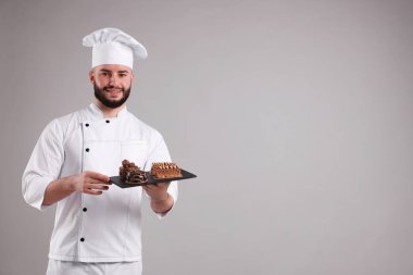 Üniformalı mutlu pastacı açık gri arka planda lezzetli çikolatalı tatlılar tutuyor, mesaj için yer var.