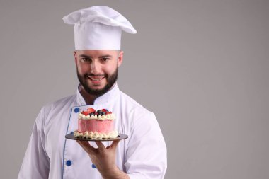 Üniformalı mutlu pastacı elinde açık gri arka planda böğürtlenli lezzetli pasta tutuyor