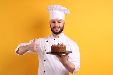 Üniformalı mutlu pastacı elinde çikolatalı pasta ve turuncu arka planda baş parmak gösteriyor.