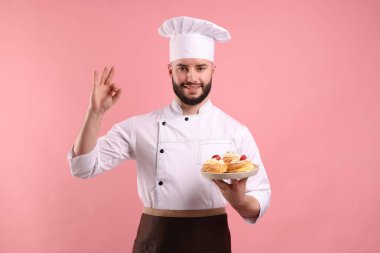 Üniformalı mutlu pastacı elinde çilekli profiterol tutuyor ve pembe arka planda güzel bir jest yapıyor.