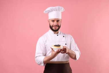 Üniformalı mutlu pastacı pembe arka planda yaban mersinli tart tutuyor.