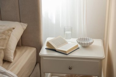 Yatağın yanında kitap, kase ve bir bardak su olan komodinin yanında.