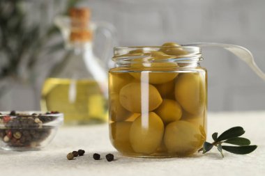 Kavanozda turşulu yeşil zeytin, çatal ve biberler beyaz desenli masada, yakın plan.