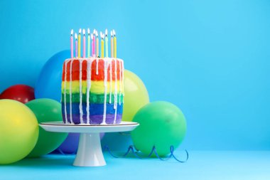 Lezzetli gökkuşağı doğum günü pastası yanan mumlar ve açık mavi arka planda balonlar, metin için alan