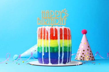 Lezzetli gökkuşağı doğum günü pastası, süslemeler ve açık mavi arka planda parti şapkaları.