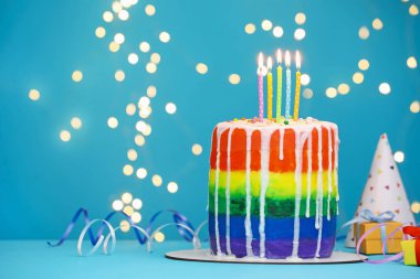 Yakıcı mumlarla, flamalarla, hediye kutularıyla ve açık mavi masada bulanık ışıklara karşı parti şapkasıyla nefis bir gökkuşağı pastası, yakın plan. Metin için boşluk