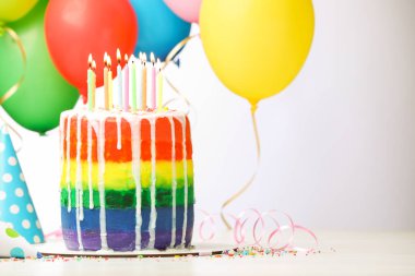 Enfes gökkuşağı doğum günü pastası, yanan mumlar, flamalar, süsler, parti şapkaları ve balonlar beyaz masada, arka planda, yakın plan. Metin için boşluk