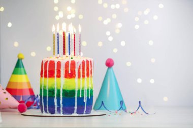 Enfes gökkuşağı doğum günü pastası, yanan mumlar, flamalar, serpiştirmeler ve parti şapkaları beyaz masanın üzerinde bulanık ışıklı arka planda, yakın plan. Metin için boşluk