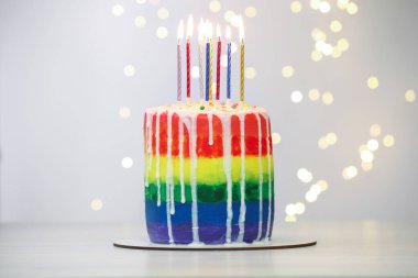 Lezzetli gökkuşağı doğum günü pastası. Beyaz masada yanan mumlar. Arka planda, bulanık ışıklar, yakın plan.