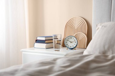 Yatağın yanında kitap, dekor, bir bardak su ve çalar saat olan komodin.