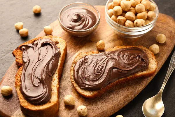 Çikolata sürülmüş ekmek dilimleri ve koyu dokulu masada fındıklar.