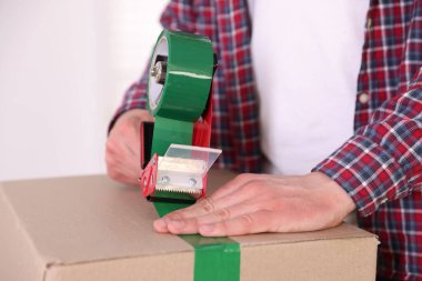 Karton kutuyu kapalı bir yere kapatmak için yapıştırıcılı bant kullanan bir adam.