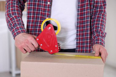 Karton kutuyu kapalı bir yere kapatmak için yapıştırıcılı bant kullanan bir adam.