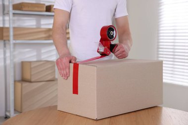 Karton kutuyu kapalı bir yere kapatmak için yapıştırıcılı bant kullanan bir adam.