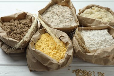 Beyaz ahşap masadaki kağıt torbalarda farklı türde un, yakın plan.