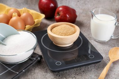 Dijital mutfak terazisi, kahverengi şeker kasesi ve gri masadaki diğer ürünler, yakın plan.