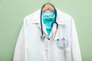 Açık yeşil arka planda asılı steteskop ve rozetli doktorun ceketi.