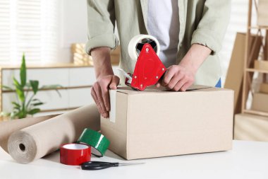 Karton kutuyu kapalı bir yere kapatmak için yapıştırıcılı bant kullanan bir adam.