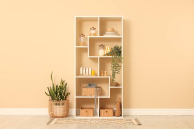 Shelving unit with dishware, jars of different products and other stuff near beige wall indoors