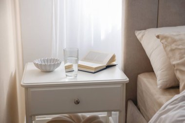 Yatağın yanında kitap, kase ve bir bardak su olan komodinin yanında.