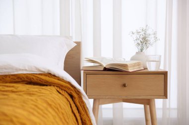 İçinde kitap olan komodin, vazoda çiçekler, yatağın yanında bir bardak su ve bardaklar.