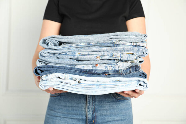 Женщина со стопкой различных стильных джинсов возле светлой стены в помещении, крупным планом