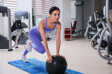 Genç bir kadın spor salonunda paspasın üzerinde ilaç topuyla egzersiz yapıyor.