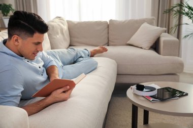 Oturma odasındaki kanepede kitap okuyan adam.