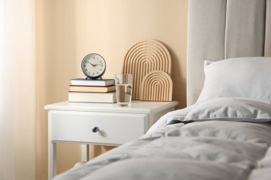 Yatağın yanında kitap, dekor, bir bardak su ve çalar saat olan komodin.