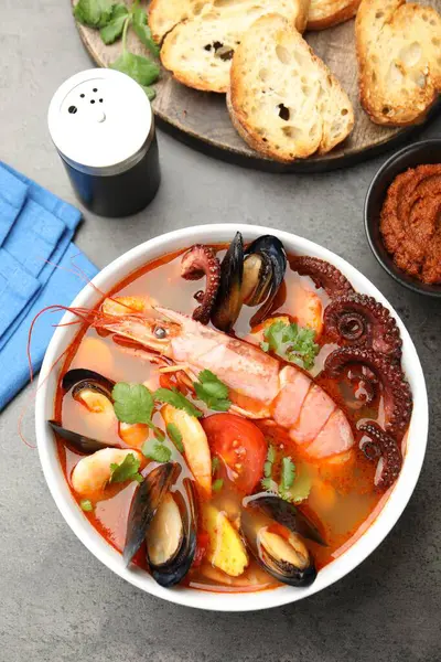 Lezzetli deniz ürünleri çorbası kasede, bir parça ekmek, sos ve baharatlar gri desenli masada, düz yerde.