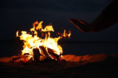 Geceleri kumsalda yanan odunların yanında elleri ısıtan bir kadın, yakın plan.