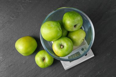 Elektronik mutfak terazisi. Koyu dokulu masada yeşil elma kasesi var.