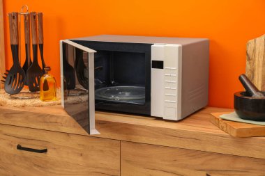 Turuncu duvarın yanındaki ahşap dolapta mikrodalga fırın ve mutfak gereçlerini aç.