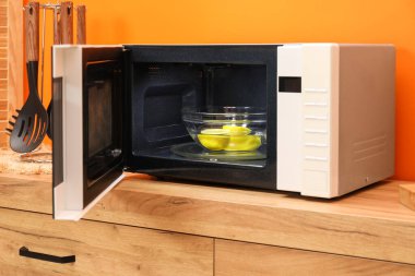 Open microwave oven with bowl of water and lemons on wooden cabinet near orange wall indoors