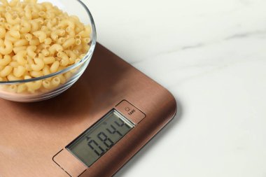Modern electronic kitchen scale with bowl of raw pasta on white marble table, closeup. Space for text