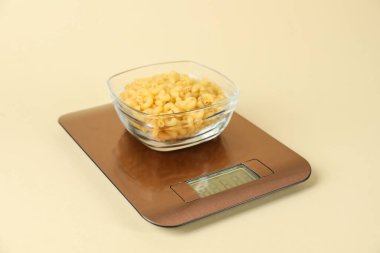 Bej arka planda bir kase çiğ makarna ile elektronik mutfak terazisi, yakın plan.