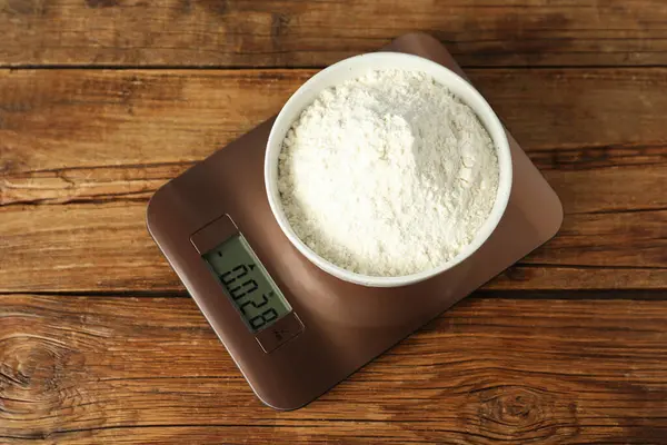 Modern electronic kitchen scale with bowl of flour on wooden table, top view