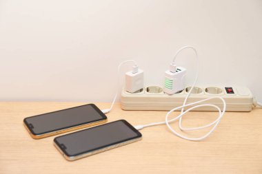 Elektrik kablosuna bağlı USB adaptörleri ışık duvarının yanındaki ahşap masada akıllı telefonları şarj ediyor.