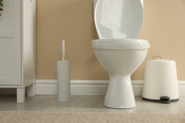 Toilet bowl, brush and trash bin near beige wall in restroom