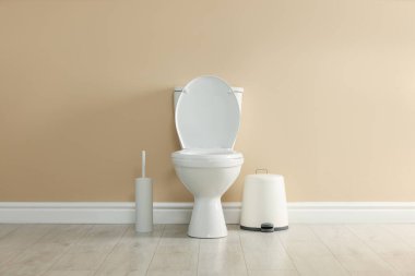 Toilet bowl, brush and trash bin near beige wall in restroom