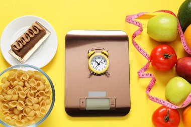 Elektronik mutfak ölçeği, alarmlı saat, ölçüm bandı ve sarı arka plandaki ürünler, düz konum. Kalori kontrolü