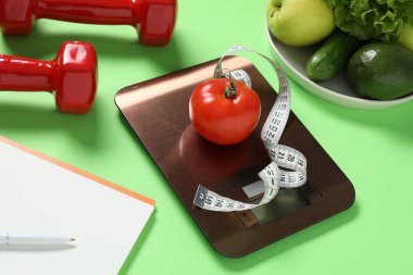 Elektronik mutfak ölçeği, mezura, dambıl, not defteri ve açık yeşil arka plandaki ürünler, yakın plan. Kalori kontrolü
