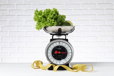 Mutfak terazisinde bir kase ürün ve tuğla duvara dayalı beyaz ahşap masada şerit var. Diyet